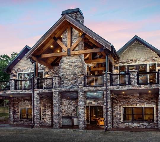 photo of timber frame home with stone facade and stone stacked supports for upper deck and fireplace. Large outdoor covered porch living space over a covered porch on lower level